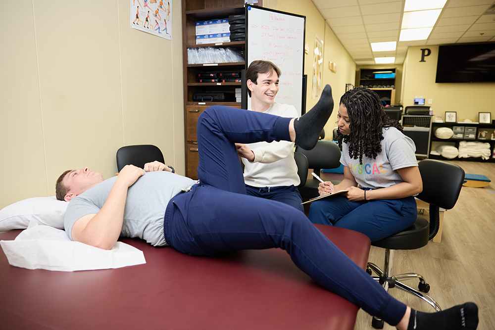 Students practice in clinic. A man lays on his back while two students examine his leg.