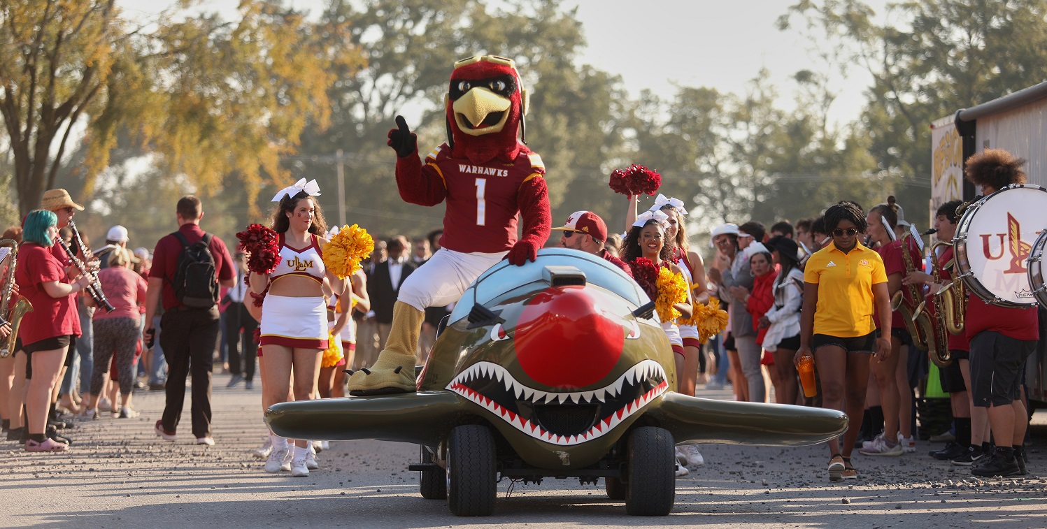 Ace the Mascot sits on a miniature P-40 airplane that is being driven on a road with both sides lined with a marching band. Cheerleaders follow him.