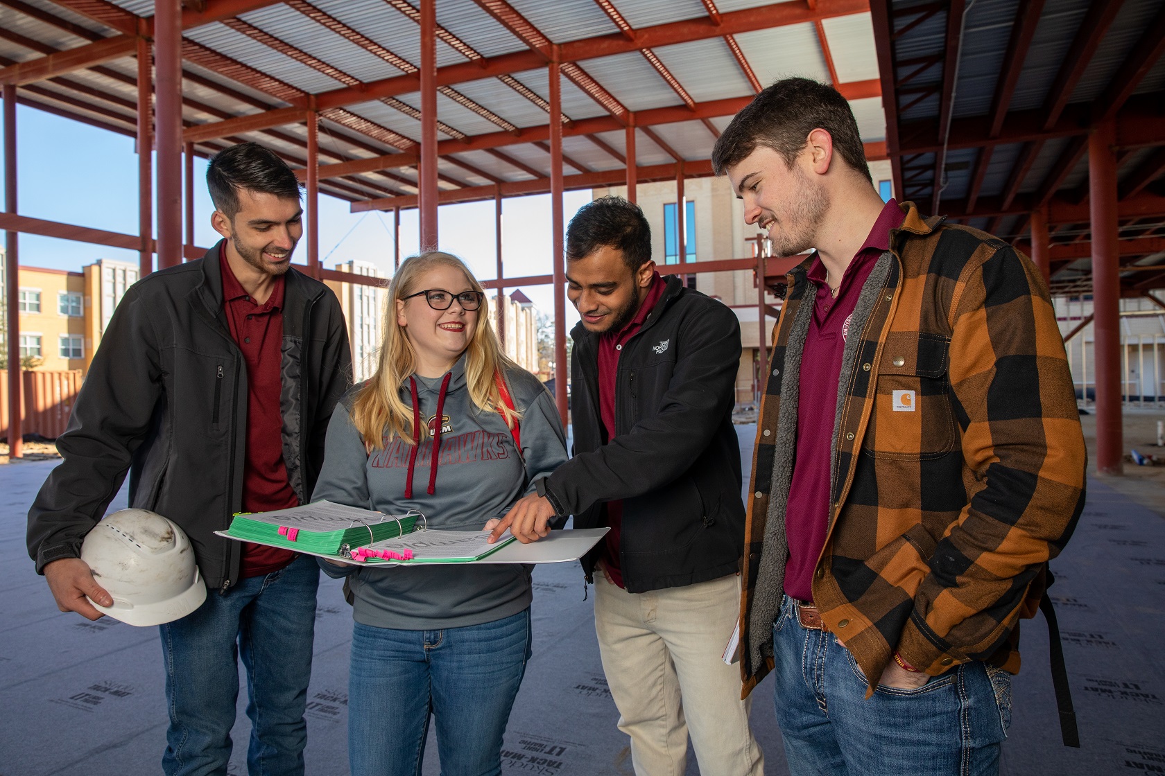 Four ULM Construction Management students stand in a construction site. They are smiling and looking at a binder.