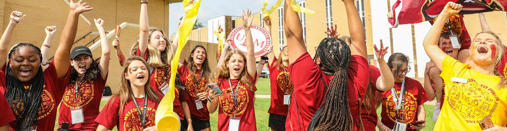 photo of about a dozen ULM students wearing matching marron and gold shirts cheer with their arms raised. With streamers and flags flying, the students exude high energy on a grassy lawn with yellow brick buildings in the background.