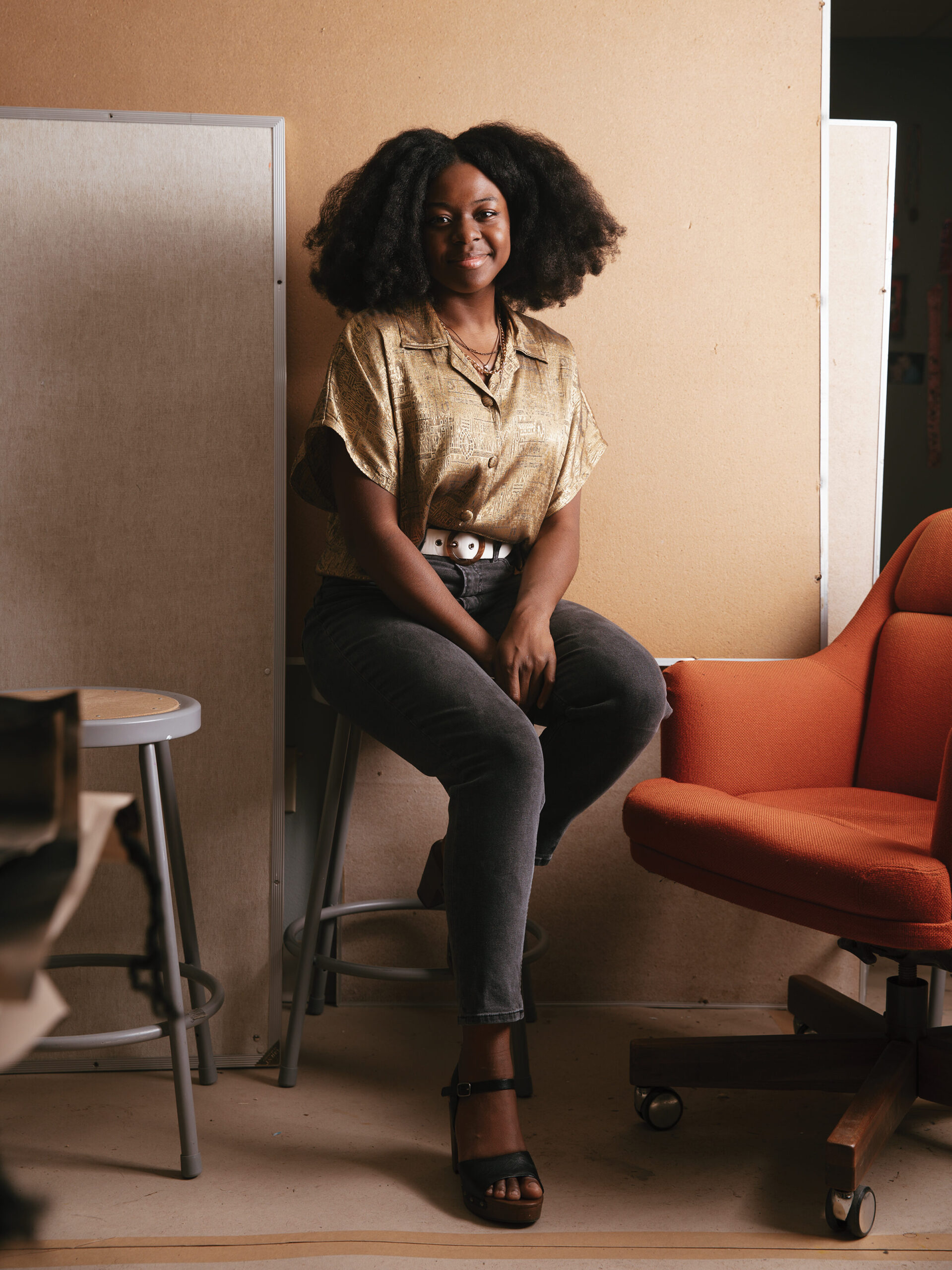 Art Professor Daija Essien is seated in front of a wall, wearing a gold shirt and gray pants.