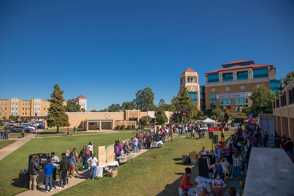 People mingle at booths and tables in the quad on ULM's campus. A banner hanging from the library in the background reads "The Best is on the Bayou."