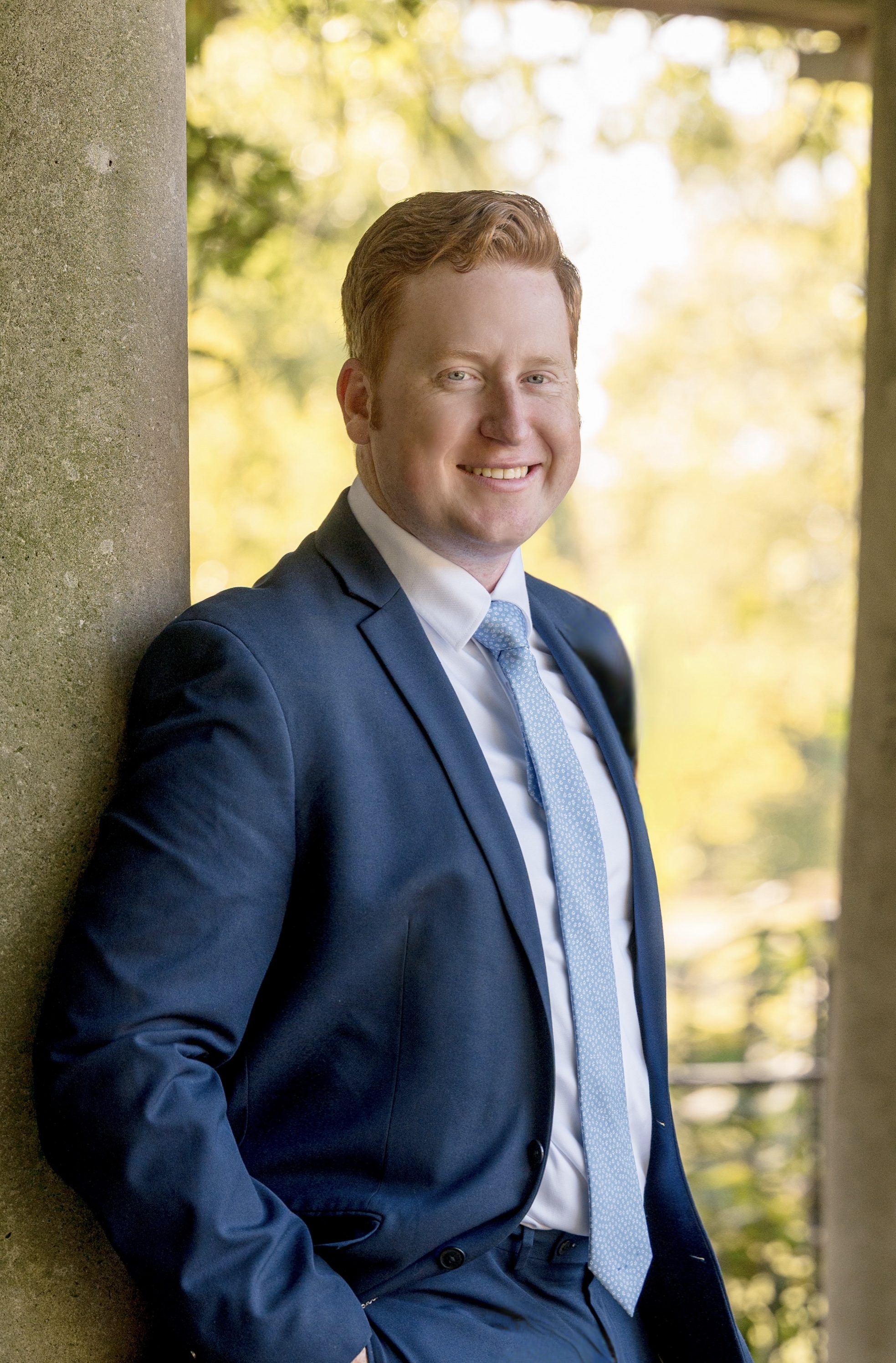 Dr. Shawn Davern is wearing a blue suit and tie and leaning against a column outside. It is a sunny day.