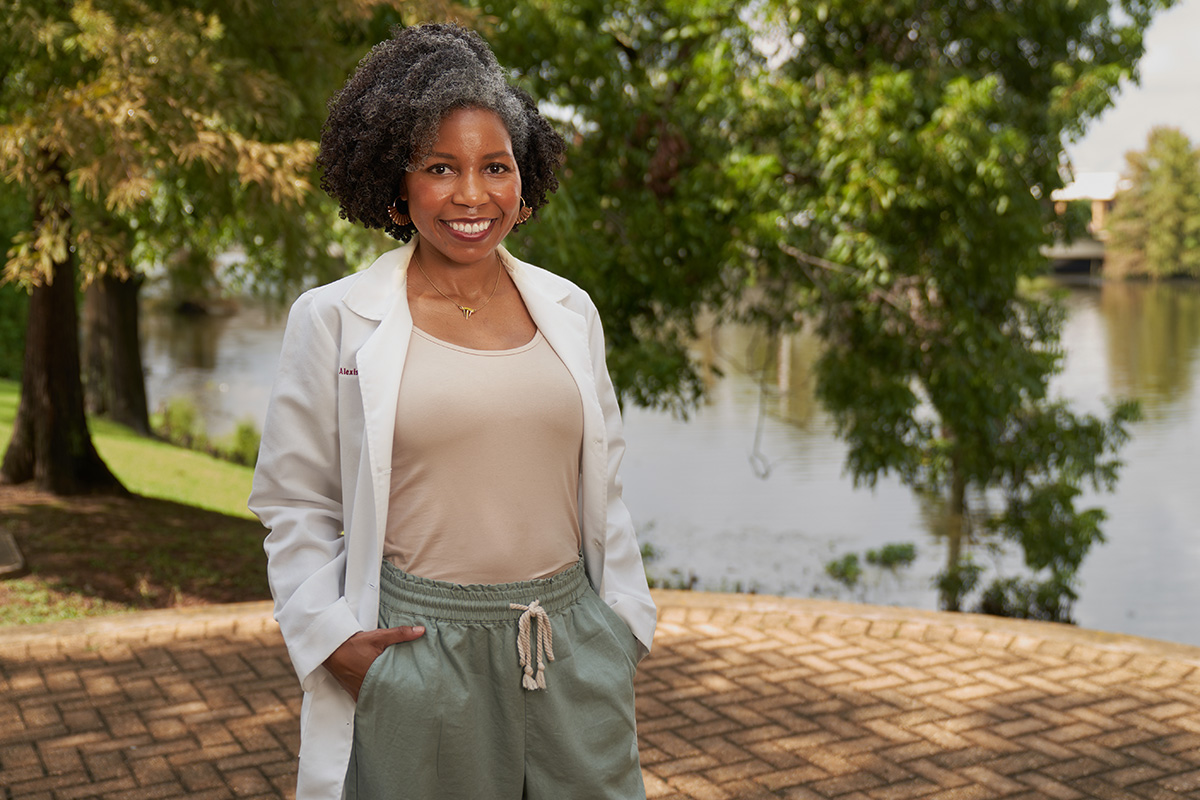A woman in a white lab coat smiles at the camera. She is standing in front of a body of water.