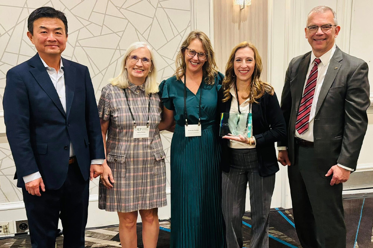 A group of two men and three women stand in a row and smile while one of the women holds and award. 
