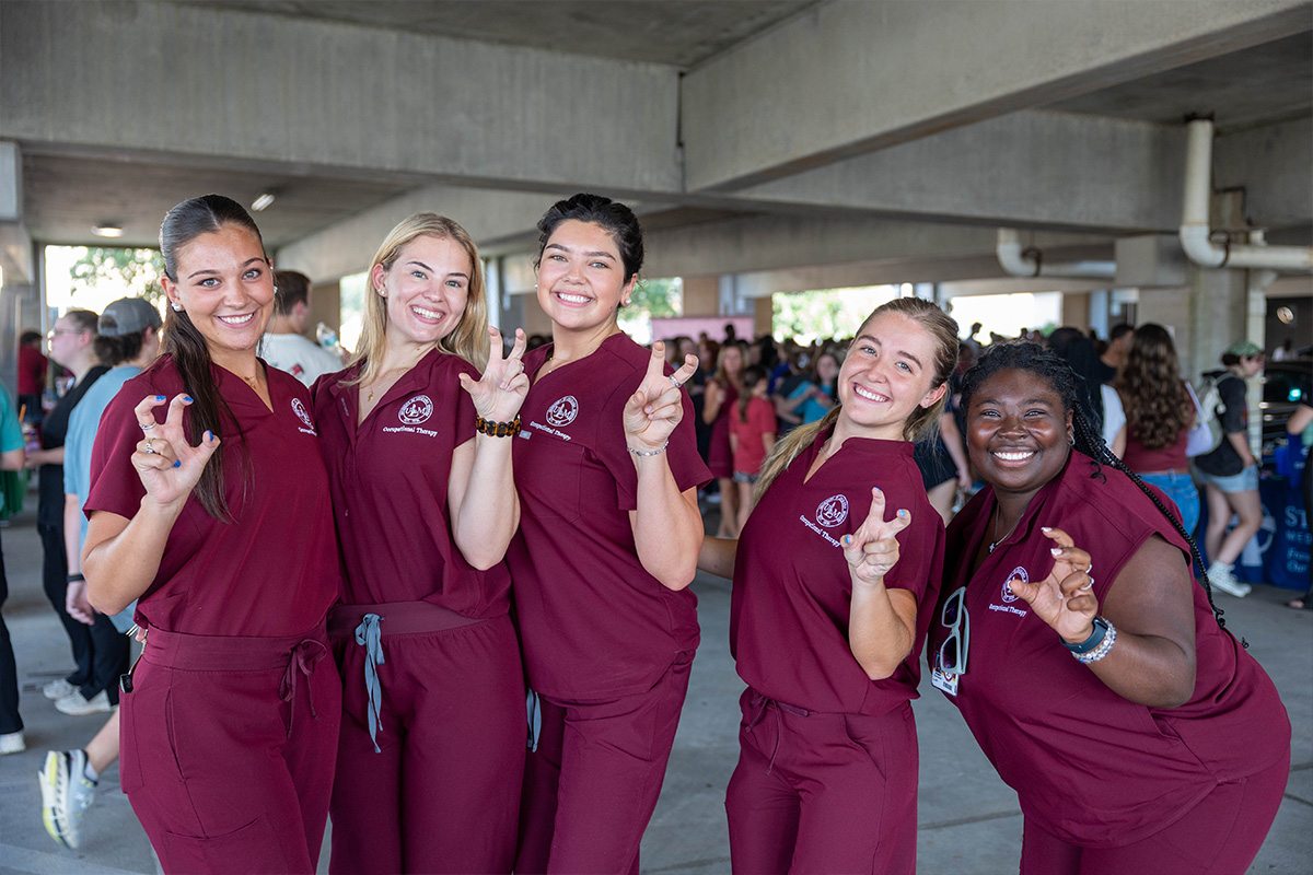 A group of female students in maroon scrubs pose at the parking garage party while making the "talons out" symbol with their hands. 