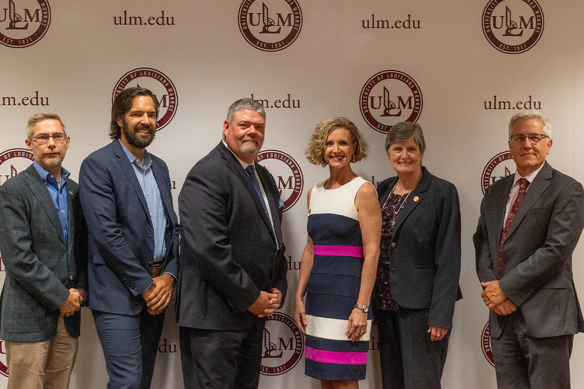 Six people stand in front of a ULM backdrop. 