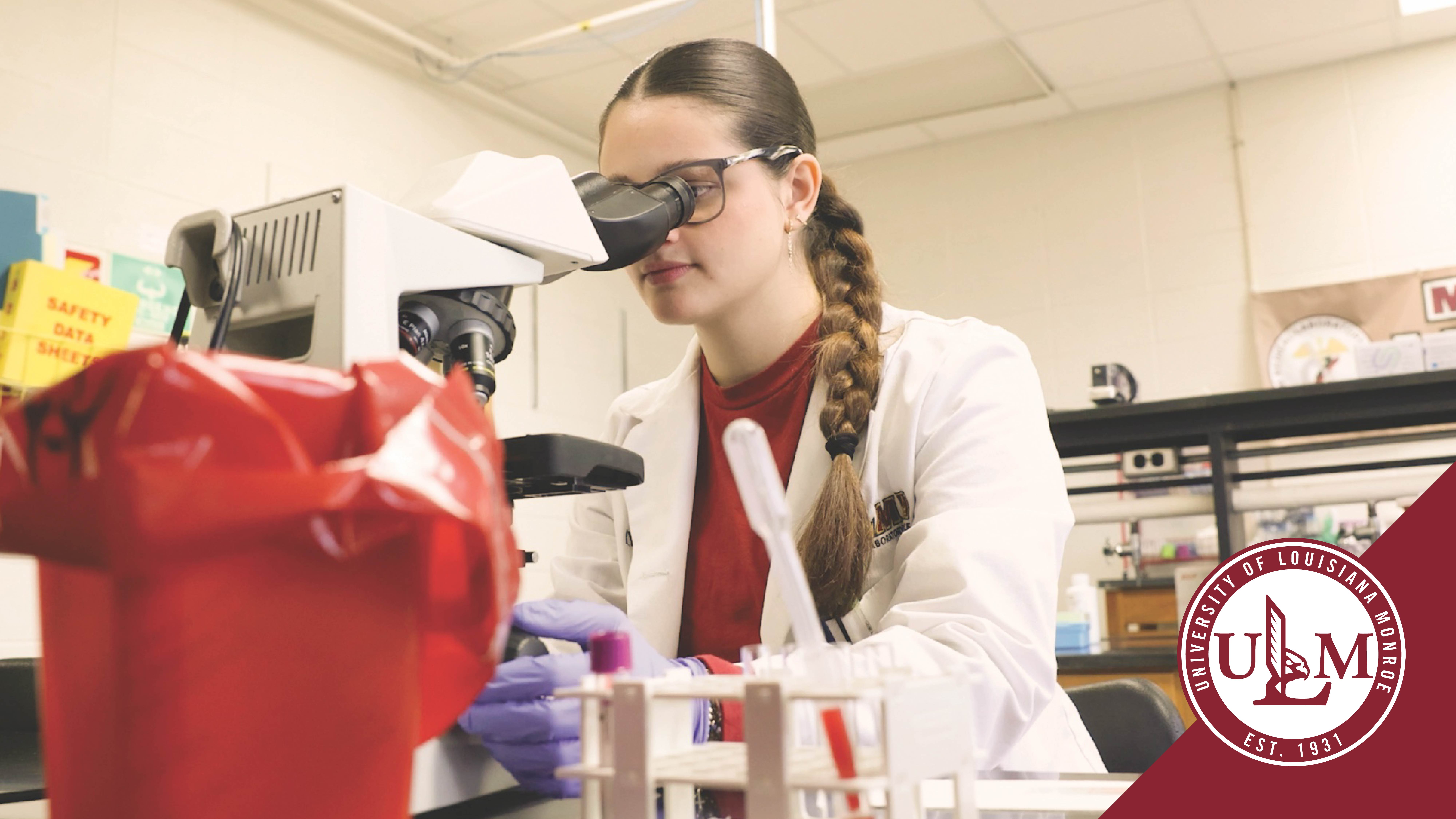 A medical laboratory science student looks into a microscope. Image has ULM logo in lower right corner.