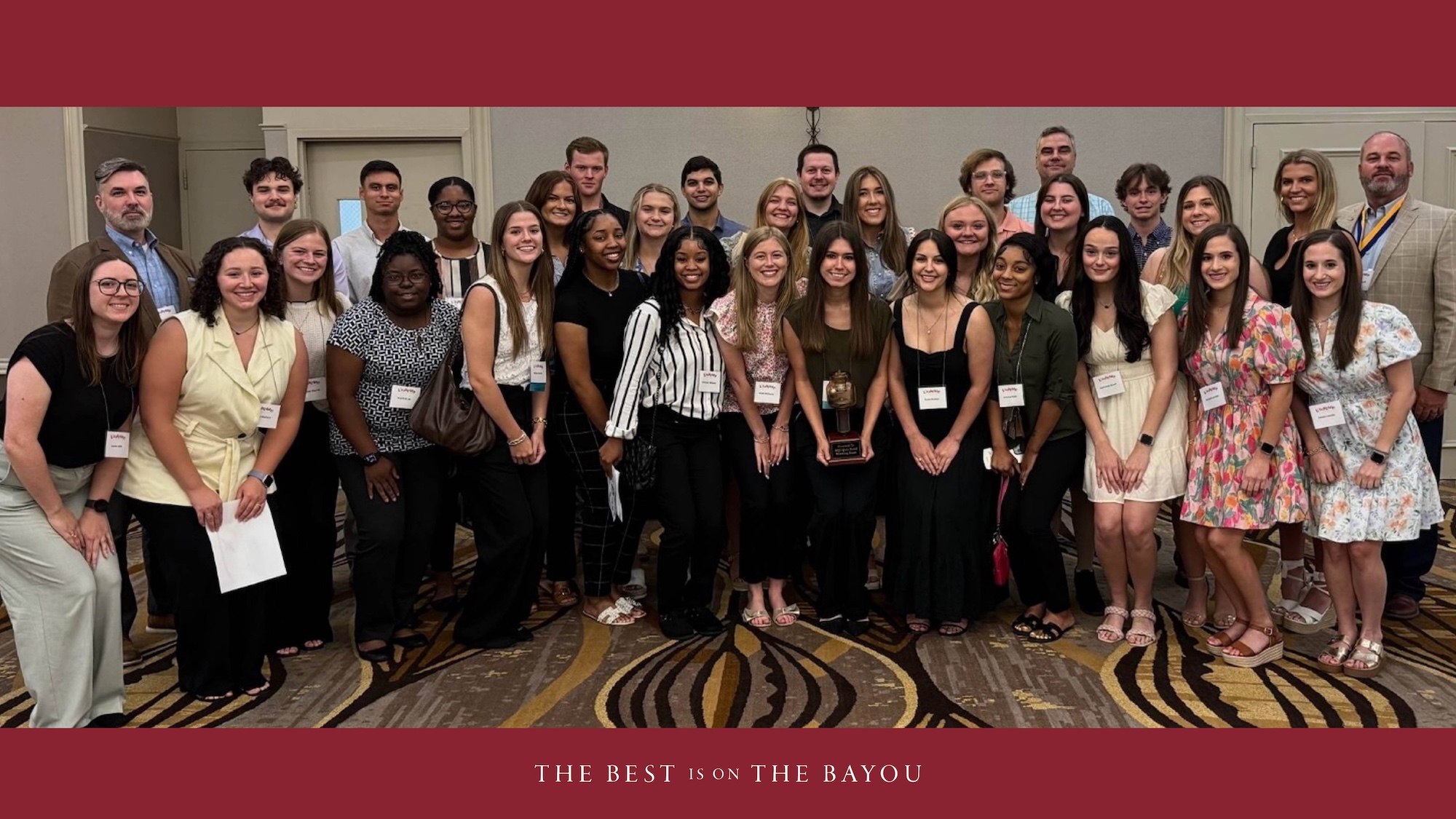 A group of students and faculty pose with smiles. The photo has a maroon top and bottom border with "The Best Is On the Bayou" at the bottom. 